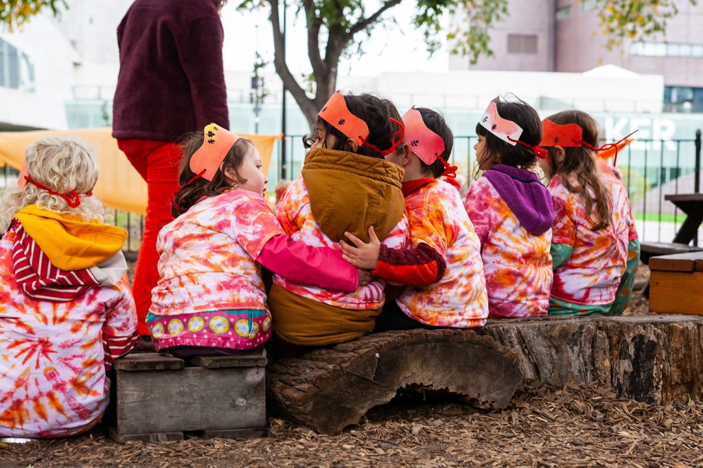 A group of kindergarten students sit together on a log, embracing each other.